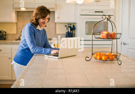 Happy mature woman typing on a laptop in her kitchen. Stock Photo