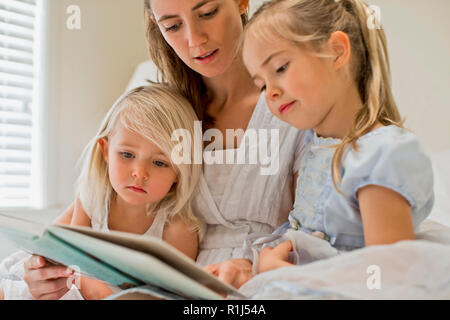 Mother sitting on a bed reading a book with her daughters. Stock Photo