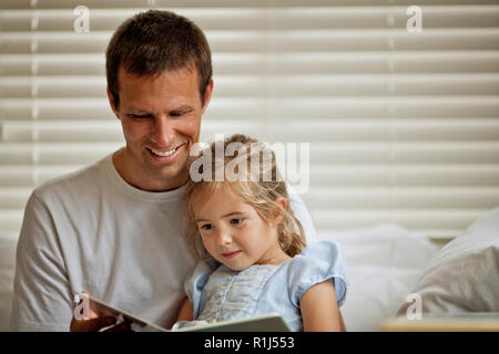 Father sitting on a bed reading a book with his daughter. Stock Photo