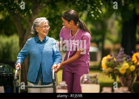 Friendly young nurse helps an elderly lady exercise with her walker in a sunny garden. Stock Photo