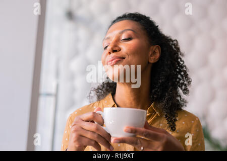 Curly dark-haired appealing woman feeling joyful while drinking coffee Stock Photo