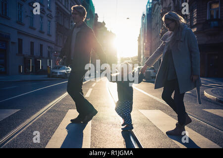 A small toddler boy with parents crossing a road outdoors in city at sunset. Stock Photo