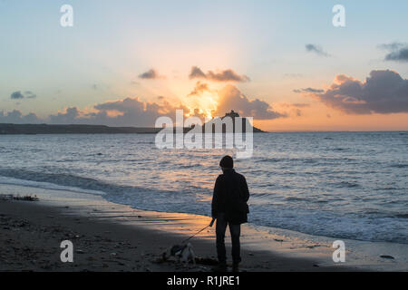 Longrock, Cornwall, UK. 13th November 2018. UK Weather. It was a mild start to the day at sunrise on the beach at Longrock. An early morning dog walker makes his way to Marazion along the beach. Credit: Simon Maycock/Alamy Live News Stock Photo