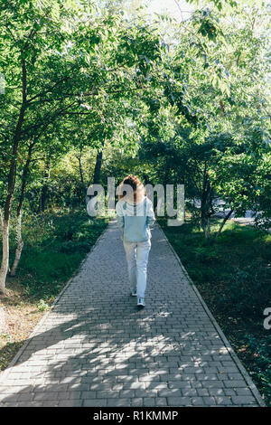 Young woman walking away on path in green park at sunny day. Full length rear view female dressed in jeans and denim jacket outdoors goes forward. Stock Photo