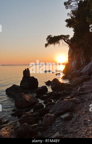 Sihouette of a young couple sitting on a rock at sunset, Kastani Mamma Mia beach, island of Skopelos, Greece Stock Photo