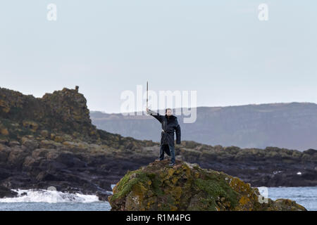 male Tourist wearing costume holding sword aloft standing on rocks on a Game of Thrones guided coach tour in Ballintoy on Northern Irelands north coas Stock Photo