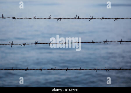 Silhouette of barbed wire fence against blurry blue water in the evening. Focused on the foreground. Stock Photo