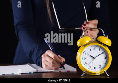 Yellow alarm old retro analog clock and girl in background writing in notes on wooden desk late at night. Time for education concept. Close up Stock Photo