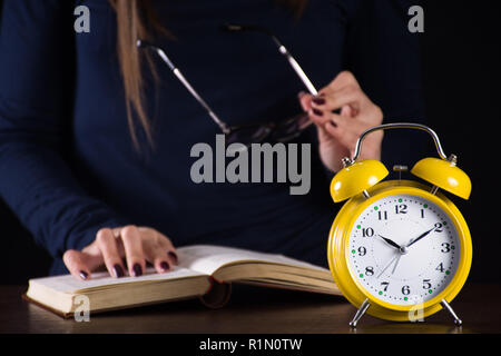 Old retro analog clock and girl in background reading book on wooden desk late at night. Time for education concept. Close up, selective focus Stock Photo