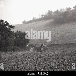 Farming in the 1950s, a farmer riding on the back of ploughing equipment being pulled by a tractor, England, UK. Stock Photo