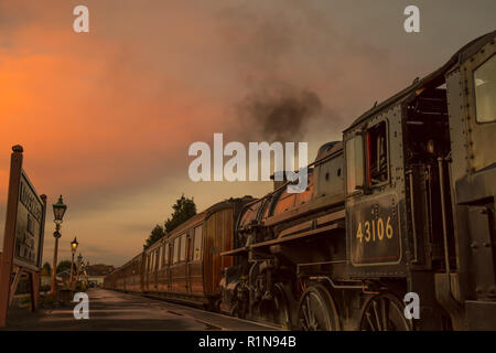 Low angle, side view close up of vintage UK steam train by platform in empty heritage railway station at end of day with glowing sunset evening sky. Stock Photo