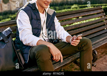 Casual happy man blogger is using a smartphone sitting on a bench in a park. Close-up Stock Photo