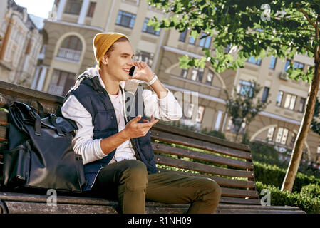 Casual happy man blogger is using a smartphone sitting on a bench in a park. Close-up Stock Photo