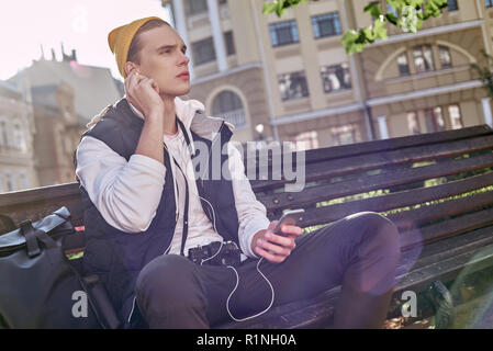 Casual happy man blogger is using a smartphone sitting on a bench in a park. He is listening music. Close-up Stock Photo