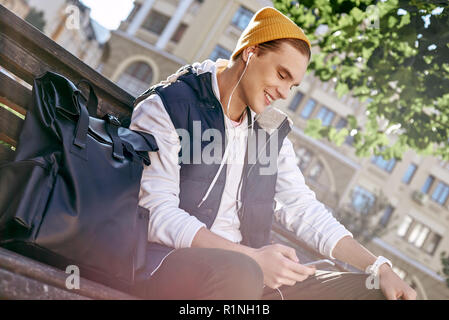 Casual happy man blogger is using a smartphone sitting on a bench in a park. He is listening music. Close-up Stock Photo