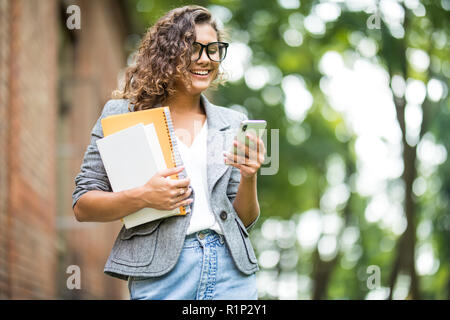 Campus life. Young smiling girl typing a message on a mobile phone while holding folders and books in a hand. Stock Photo