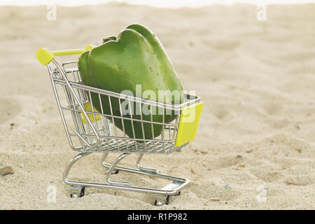 Green sweet pepper paprika in shopping trolley standingon the sand. Green bell pepper on cart for sell. Vegetable loaded in a miniature shopping cart  Stock Photo