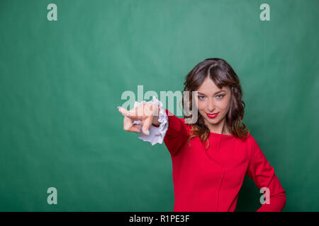 Image of cheerful woman with long brown hair winking and showing index finger aside meaning hey you isolated over green background.Emotional expressing woman in red dress, red lips and dark curly hairstyle.Copy space Stock Photo