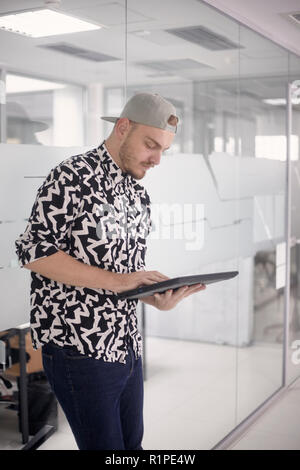 one young man standing, 20-29 years old, working in modern office space, using tablet computer. casual clothes. Stock Photo