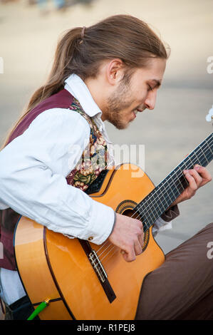 Krakow, Poland: A busker plays guitar in the old market square. Smiling, seated musician performs on an accoustic guitar to a crowd of people. Stock Photo