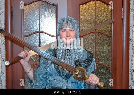 A man dressed up in a knight's hauberk at home in his room. Knight's sword in the hands of a man. Stock Photo