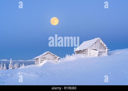 Winter landscape with old wooden hut in a mountain village. Morning twilight with a full moon and clear blue sky. Fresh snow and frost on the walls an Stock Photo