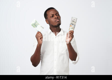 Young attractive african man holding USA currency and euro currency deciding which one to choose. How is it better to save money concept Stock Photo