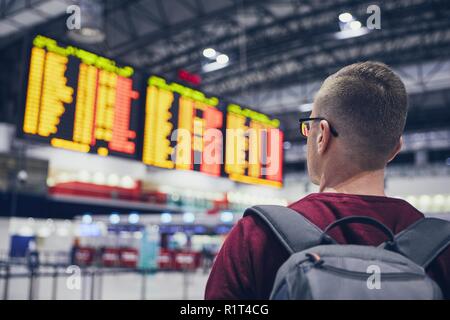 Rear view of young man checking flight schedule on the arrival departure board at airport. Stock Photo