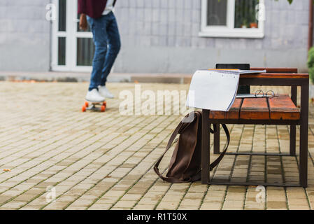 cropped shot of student riding skateboard with backpack and notebook on bench on foreground Stock Photo