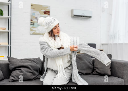 freezed young woman putting on warm clothes while sitting on couch Stock Photo