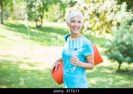 Portrait of senior woman holds fitness mat on her back in the park and preparing for exercise Stock Photo