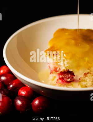 Slice of cranberry cake in shallow white bowl drizzled with lemon sauce.  Portrait crop on black background.  Fresh cranberries in foreground. Stock Photo