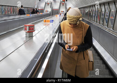 Tube commuters time, Young woman wearing a Hijab and using her smartphone as she travels up the escalator on the London Underground Stock Photo
