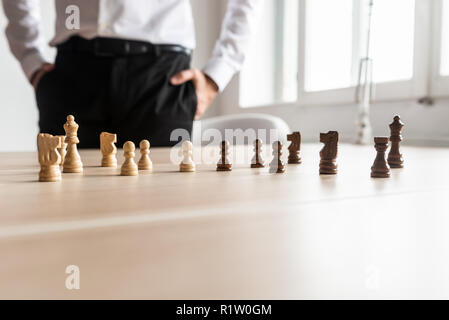 Businessman standing at his office desk looking at black and white chess pieces arranged on the wooden desk. With copy space on the lower part of the  Stock Photo