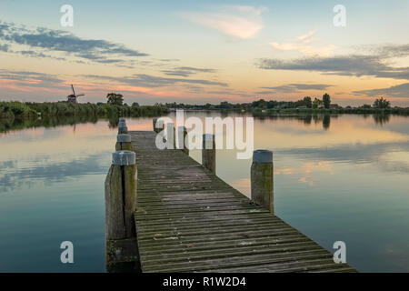 Wooden pier a the river Rotte at sunset, close to Rotterdam in The Netherlands. In the distance a typical dutch windmill can be seen. Stock Photo