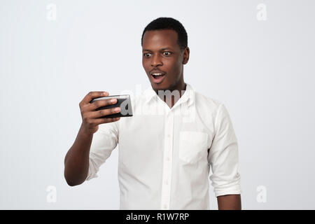 African man in white shirt playing mobile game with shocked face. Stock Photo