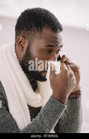 cold african american man in warm scarf with runny nose holding napkin while talking on smartphone Stock Photo