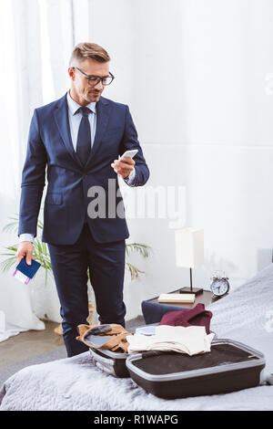 smiling businessman in suit and eyeglasses holding passport with boarding pass and using smartphone while packing suitcase at home Stock Photo