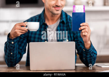 cropped shot of smiling man holding cup of coffee and passport with boarding pass while using laptop at home Stock Photo