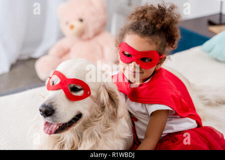Smiling adorable little african american kid with happy dog in red superhero mask Stock Photo