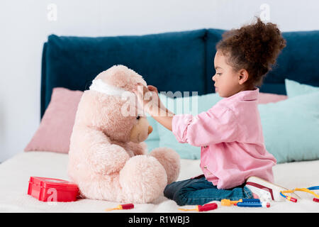 Little african american child in pink jacket playing the doctor  with teddy bear Stock Photo