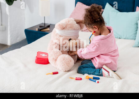 Little african american child in pink jacket playing the doctor  with teddy bear Stock Photo