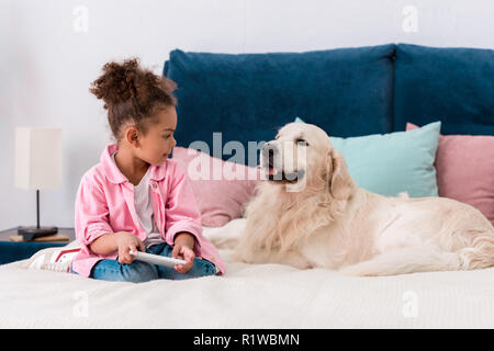 cute african american child sitting on the bed with golden retriever and playing with smartphone Stock Photo
