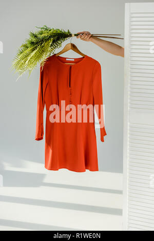 female hand holding hanger with red stylish dress and green plants near room divider Stock Photo