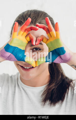 young transgender man looking at camera while making heart sign with hands in colors of pride flag in front of white brick wall Stock Photo