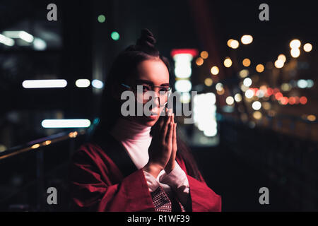 beautiful asian girl in burgundy kimono making namaste gesture on street with neon light in evening, city of future concept Stock Photo