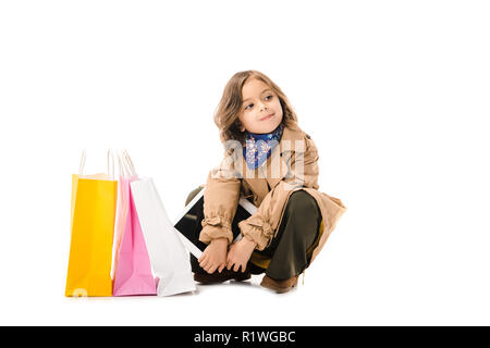 beautiful little child in trench coat sitting on floor with colorful shopping bags isolated on white Stock Photo