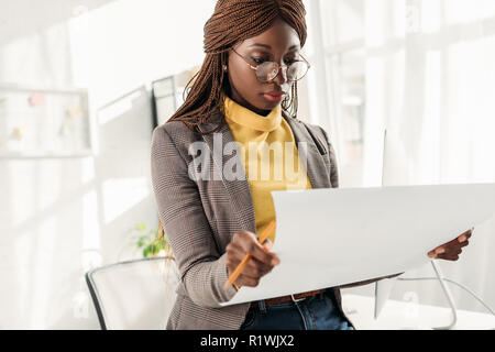 focused african american female architect in glasses holding blueprint and pencil in office Stock Photo
