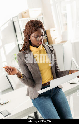 concentrated african american female architect in glasses holding blueprint and pencil in office Stock Photo