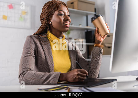 smiling adult african american adult businesswoman sitting at computer desk, drinking coffee and using graphic tablet Stock Photo